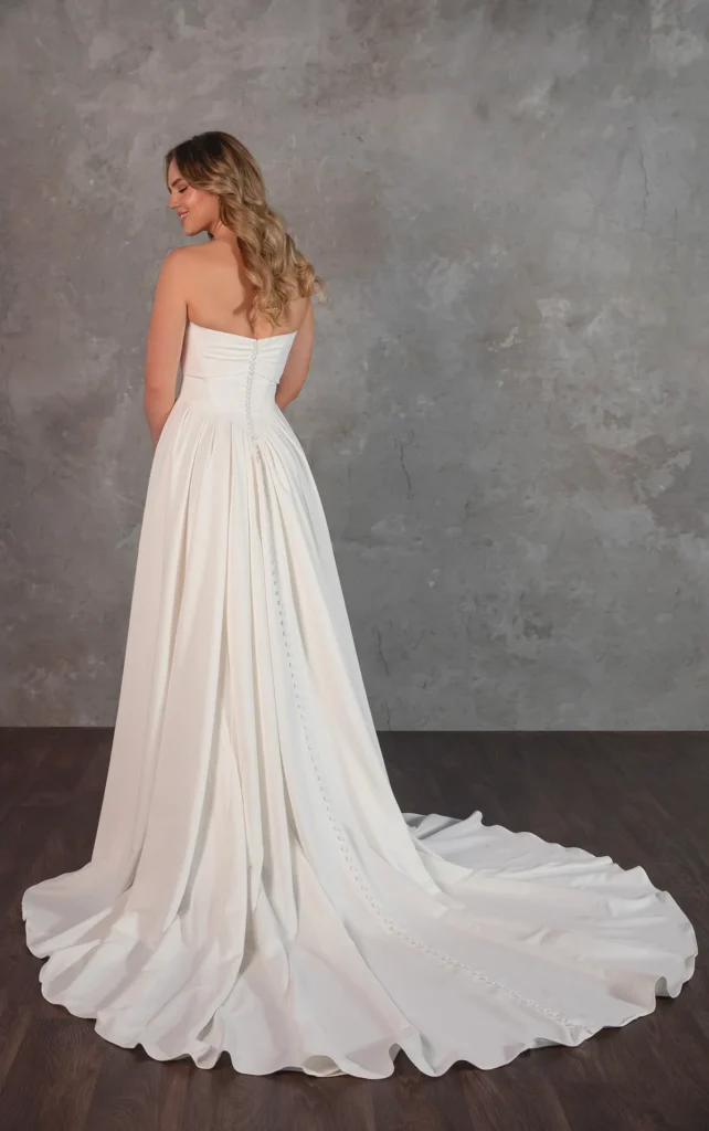 Effortless elegance meets Greek goddess inspired beauty a minimalist matte satin wedding gown designed for the modern bride. The strapless bodice features soft, draped crisscross detailing that flatters the figure, creating a look that is both timeless and sophisticated. From the structured Basque waist, the classic A-line skirt flows gracefully over the hips, draping beautifully with every step. This chic wedding dress is a fusion of simplicity and refinement. This gown is also available in plus sizes.