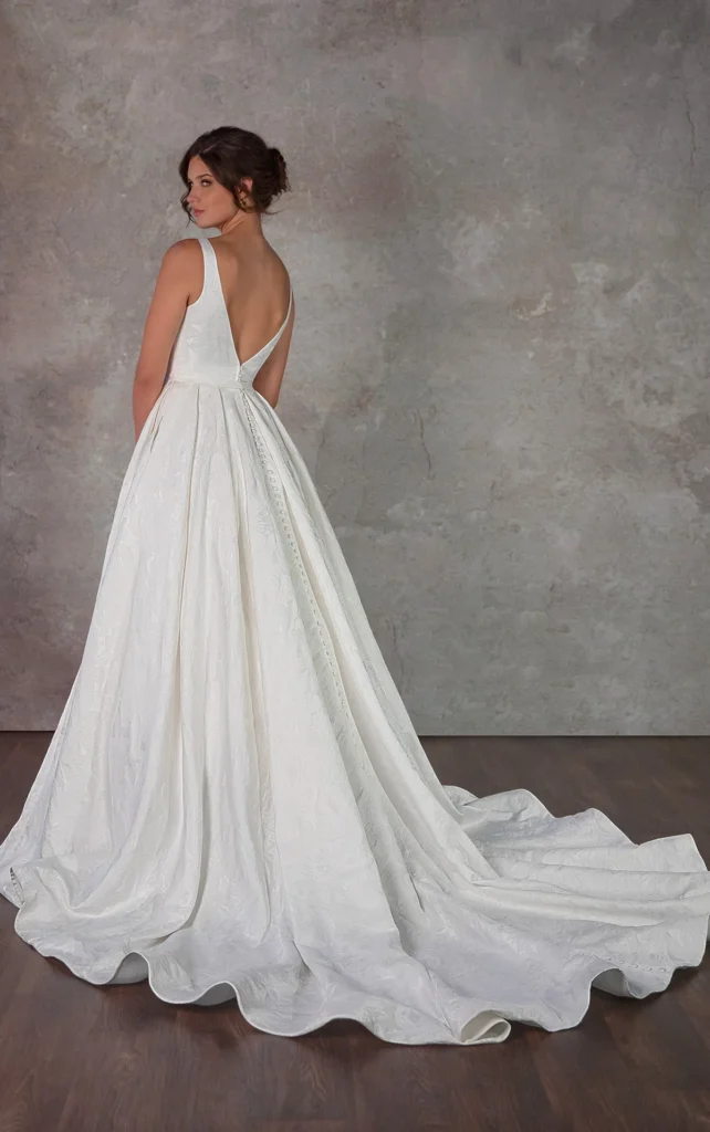 A modern bride’s dream comes to life in Candy, a stunning textured satin wedding dress that eschews traditional lace for contemporary elegance. The A-line silhouette begins with a daring deep V-neckline, plunging from sleek shoulder straps to create a sculpted, contoured bodice. Lined with illusion tulle for modesty and comfort, this sexy A-line wedding gown balances allure with sleek sophistication. A matching V-back detail mirrors the front, while a subtle waistband marks the transition to a full, pleated skirt where the luxurious textured satin truly shines. Discreet side pockets add a practical touch to this sleek bridal look, available in timeless ivory or showstopping gold for the fashion-forward bride. This gown is also available in plus sizes.