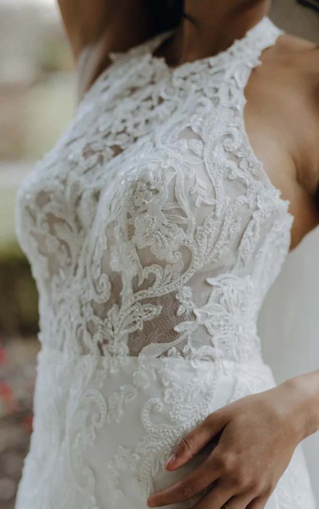 Sheer romance comes to life in Harriett a sexy lace halter neck wedding gown designed with the modern bride in mind. A high halter neckline wraps around the neck, forming an eye-catching graphic lace bodice and open back detail that reveals the corset structure underneath. A discreet row of fabric-covered buttons line the center back to resolve at the hip where modest ruching and expertly-placed lace appliqués flatter the form and accentuate the natural curves of the body. Past the hip, this romantic fit-and-flare bridal gown flares into a soft tulle sweeping train scattered and shaped by floral lace accents for a final enchanting detail.