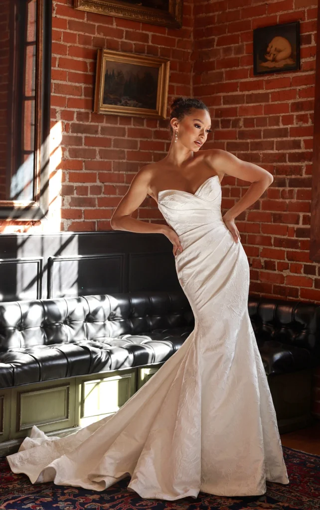 Timeless elegance meets modern sophistication in Jodie, a stunning strapless column wedding dress designed for the bride who loves both classic and contemporary style. Crafted from luxurious satin jacquard or satin, the structured bodice features a draped sweetheart neckline with delicate ruching that enhances the figure. The sleek column silhouette is complemented by a detachable pleated overskirt, adding dramatic movement and versatility. A row of fabric-covered buttons trails down the back for a vintage-inspired touch, blending refined glamour with effortless romance. This gown is also available in plus sizes.