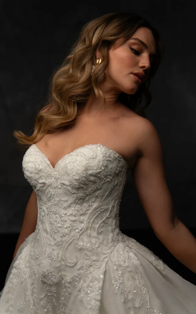 Classic bridal elegance meets modern glamour in this breath taking ballgown. Samara features a beautifully embellished sweetheart neckline highlighting the decolletage, adding a touch of romance to the structured bodice. The grand silhouette comes to life with shimmering glitter tulle, while pleated layers create soft movement and dimension. Delicate scrolls of lace cascade throughout, adding a regal yet ethereal touch. For a timeless finish, fabric-covered buttons trace from the top of the neckline down to the edge of the sweeping train. This showstopping gown is also available in plus sizes.